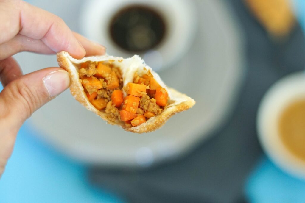 Gyoza mit Hack-Füllung im Anschnitt, sodass man sowohl die Karottenwürfel als auch das Hack gut sieht. Eine Hand hält die frittierte Teigtasche. Im Hintergrund sieht man unscharf Sojasoße.
