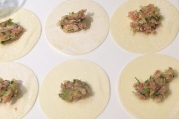 Teigtaschen füllen: Sechs Dumpling-Wrapper liegen auf hellem Hintergrund. Sie sind jeweils mit einer japanischen Garnelen-Gemüse-Mischung belegt und bereit, um gefaltet zu werden.